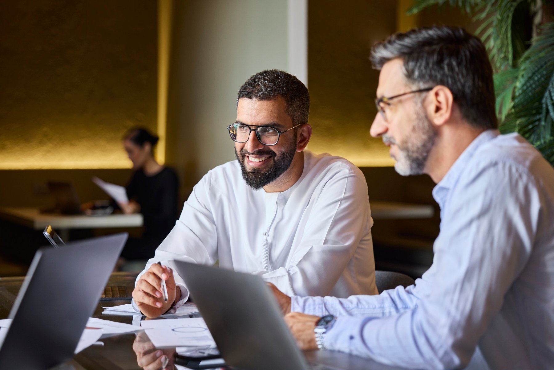 Smiling male entrepreneur sitting next to colleague at coworking space. Businessmen are communicating in meeting. They are wearing eyeglasses.