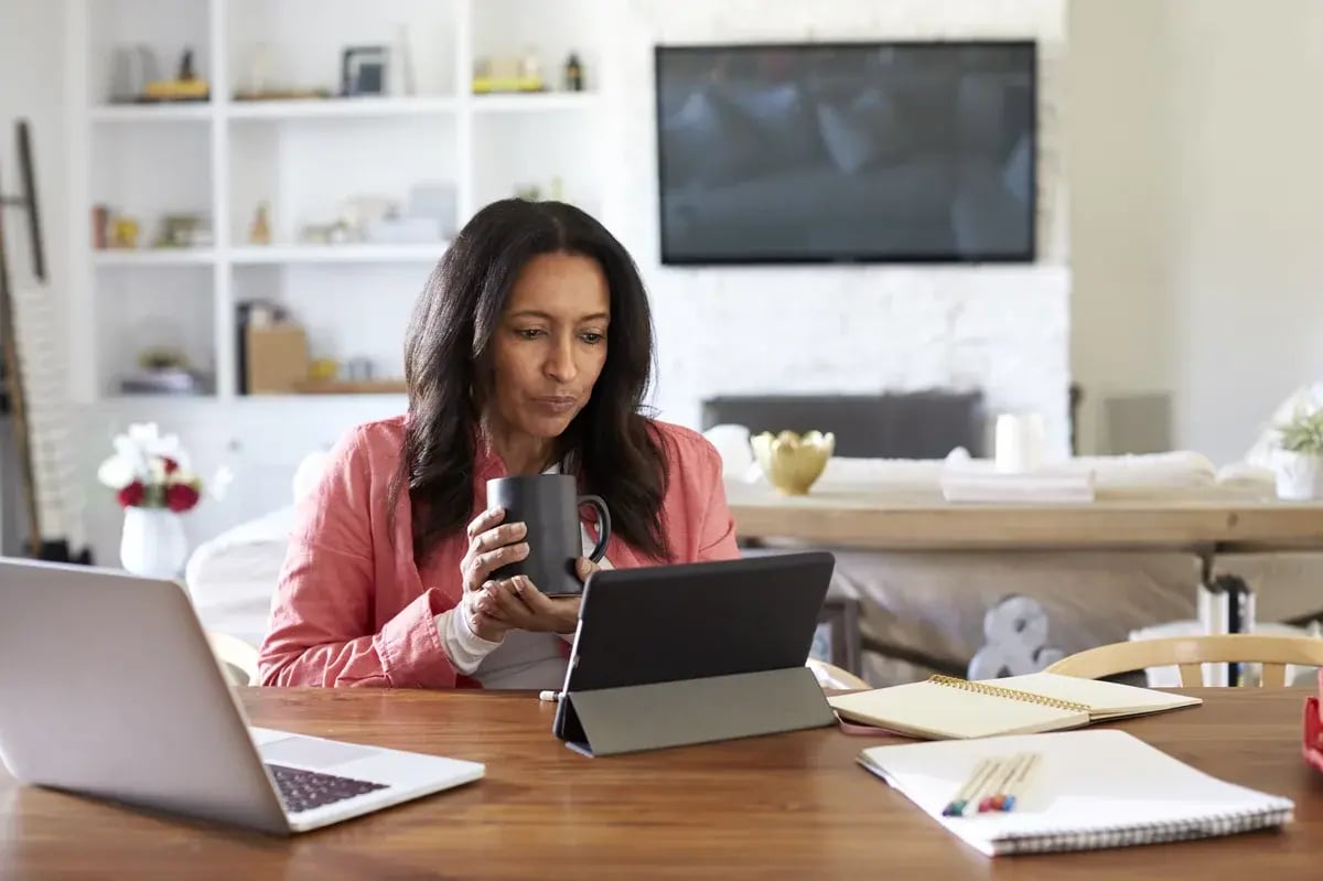 Middle aged woman sitting at a table reading using a tablet computer, holding a cup, front view Middle aged woman sitting at a table reading using a tablet computer, holding a cup, front view
